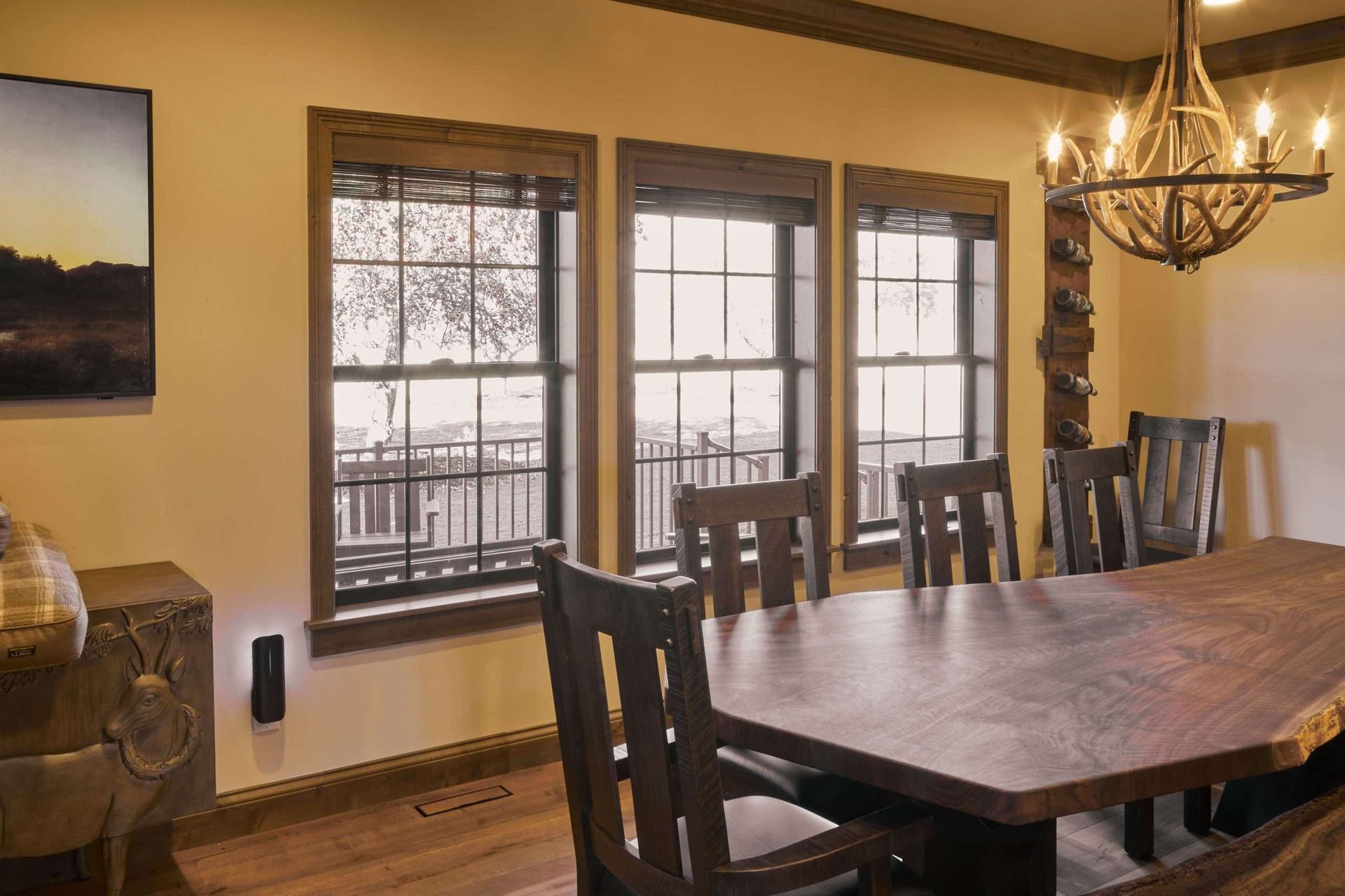 Dining room with wooden table, chairs, chandelier, and large windows overlooking a deck.