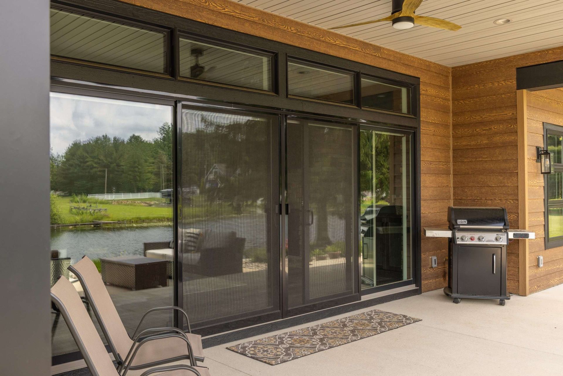 Covered patio with sliding glass doors, lounge chairs, and a grill beside a wooden wall