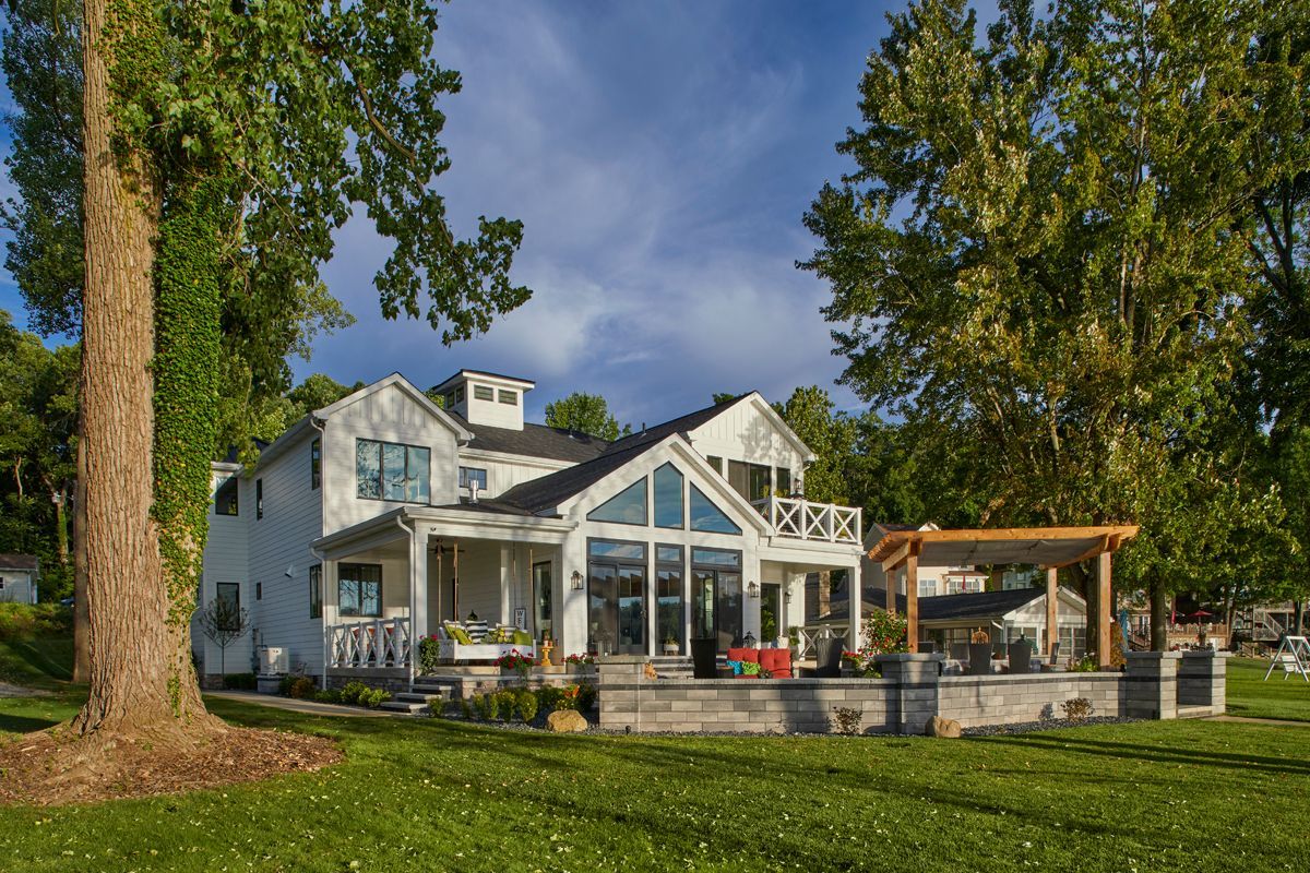 White lakeside house with large windows, a deck, and a green lawn under tall trees
