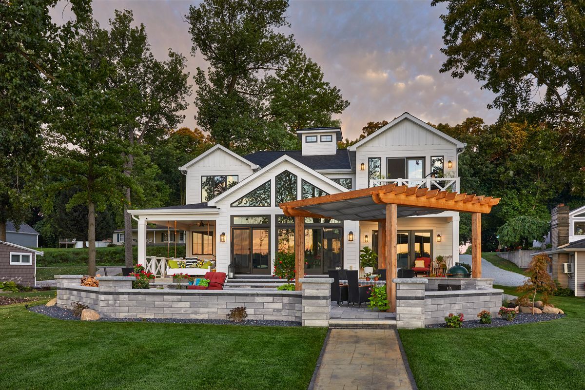 House with a wraparound porch and pergola at dusk, framed by trees and a front walkway.