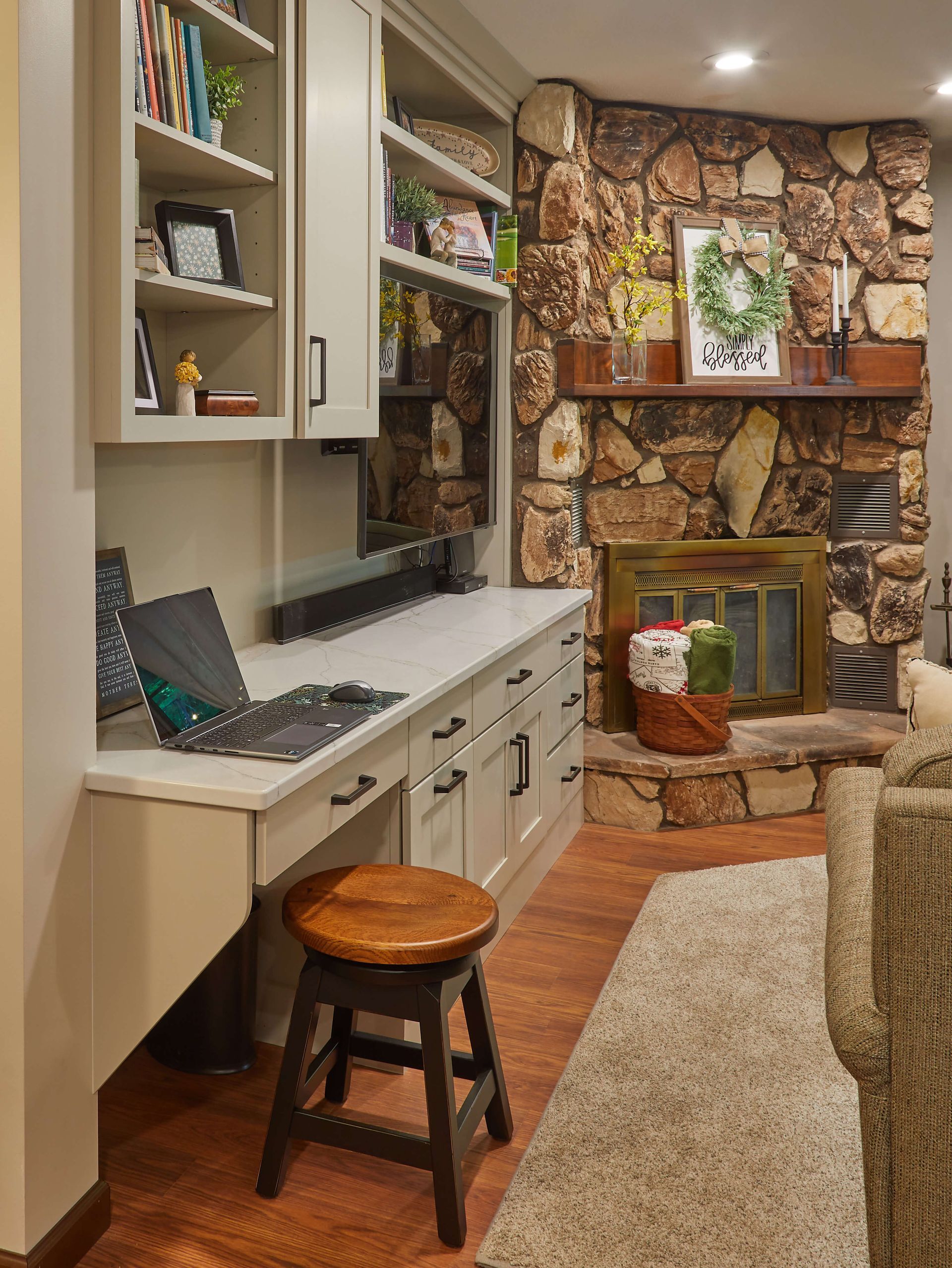 Cozy home office with built-in desk, laptop, stool, stone fireplace, and shelving