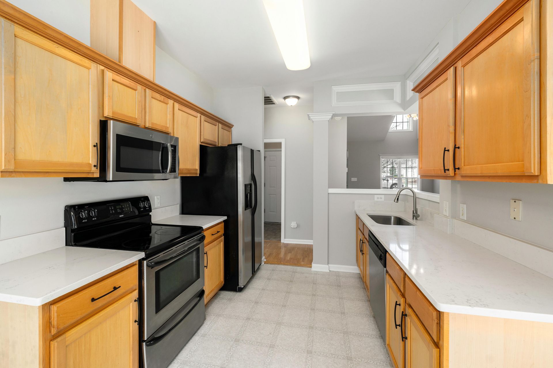 Bright kitchen with light wood cabinets, black appliances, and white countertops around an empty walkway.
