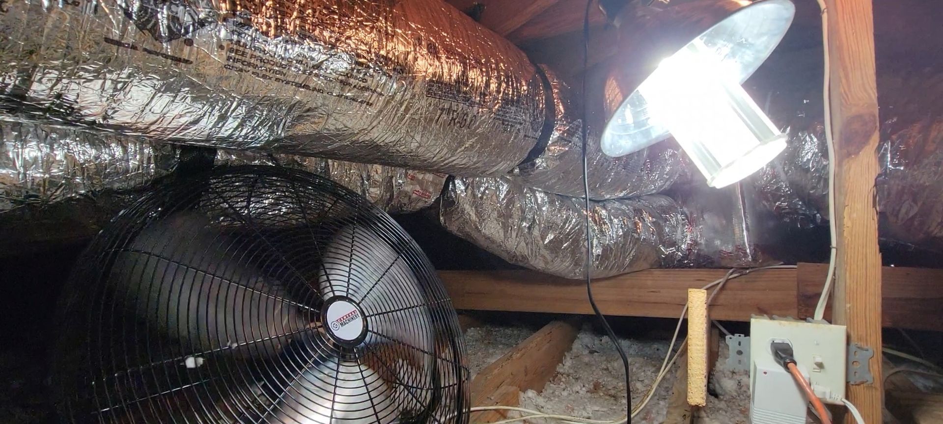A fan is sitting in an attic next to a lamp.