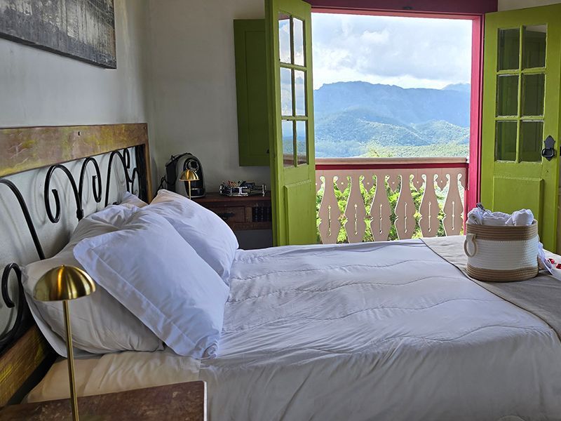 Quarto luminoso com janela aberta, persianas verdes e vista para as montanhas, além de varanda.