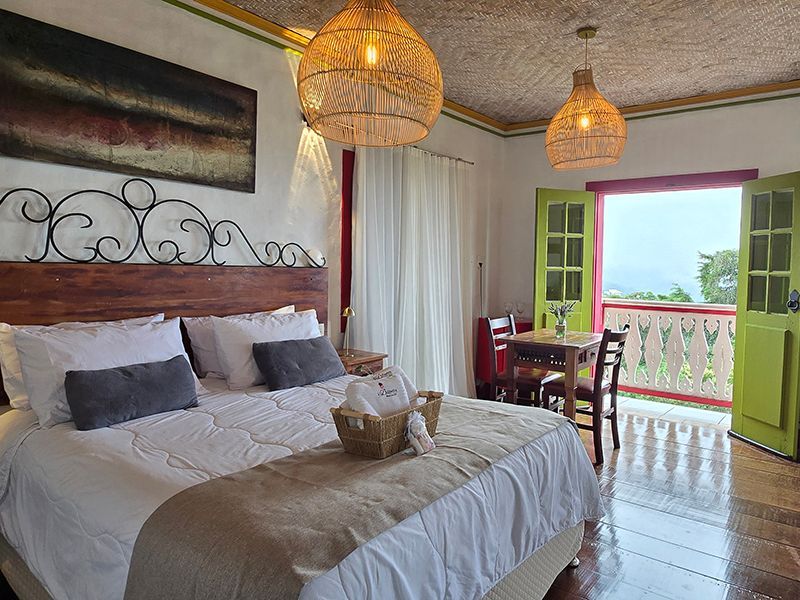 Quarto luminoso com cama, candeeiros de vime e porta de varanda aberta com vista para o mar.