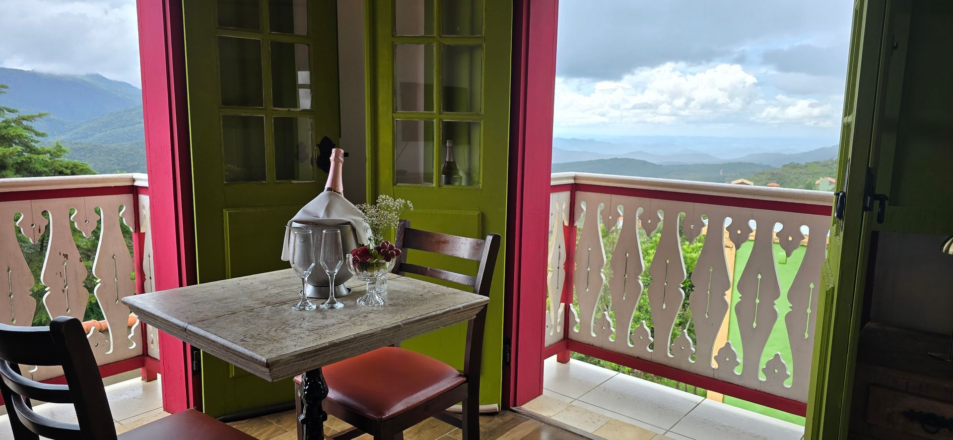 Pátio aconchegante com mesa de madeira, cadeiras e portas abertas com vista para as montanhas e a vegetação.