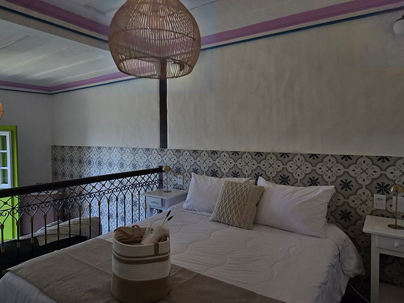Cama com lençóis brancos e luminária pendente de vime em um quarto ensolarado com piso de azulejo.