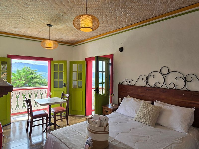 Quarto luminoso com cama ornamentada, portas francesas abertas, cadeiras de varanda e detalhes coloridos com vista para a vegetação.