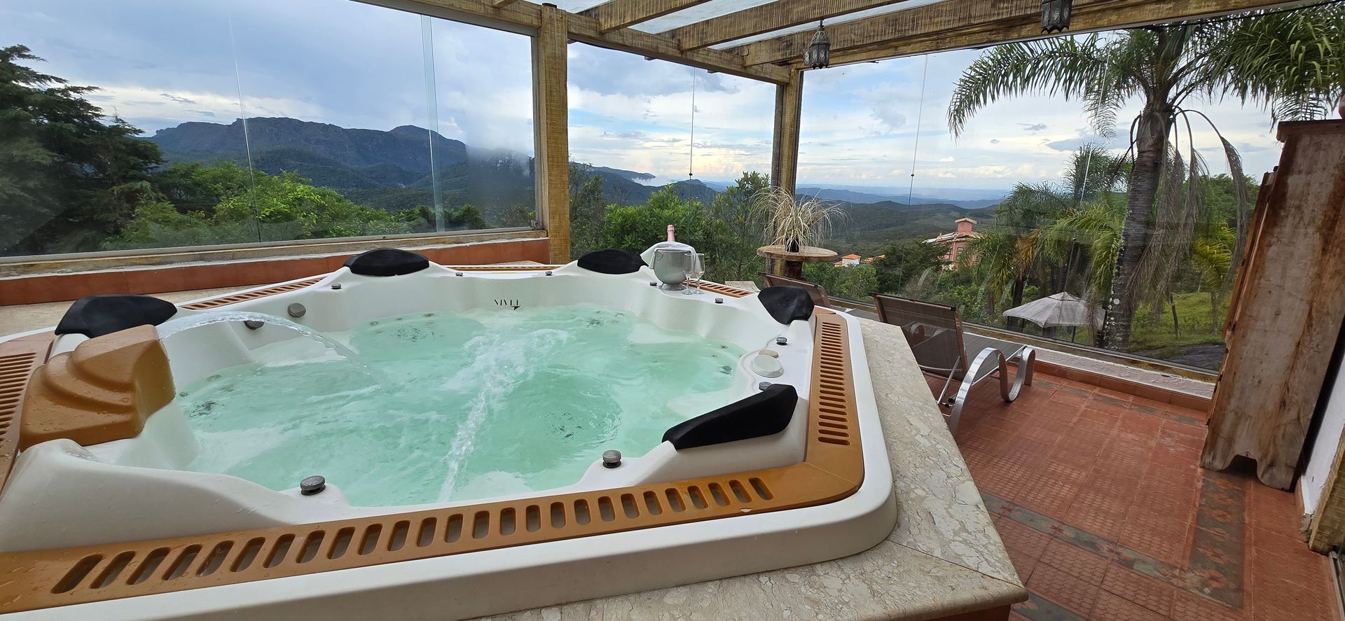 Banheira de hidromassagem ao ar livre em um terraço com vista para as montanhas ao fundo, rodeada por espreguiçadeiras e vegetação.