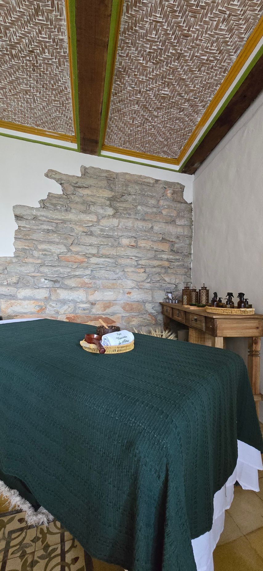 Quarto pequeno com cama coberta em verde escuro, uma parede com detalhes em pedra e uma cômoda de madeira com garrafas.