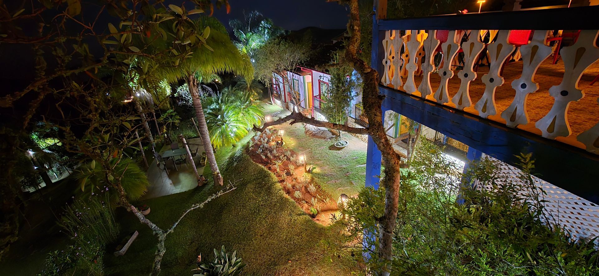 Caminho noturno em um jardim ao lado de uma ponte ou varanda iluminada, com árvores, flores e luzes coloridas iluminando a cena.