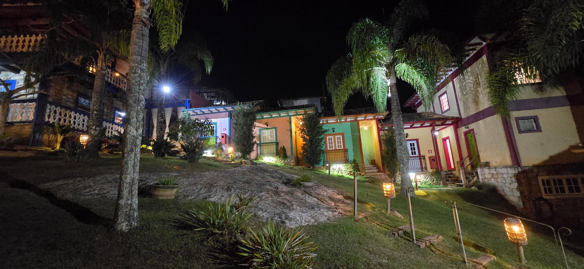 Vista noturna de um pátio colorido com cabanas iluminadas, árvores e um caminho pavimentado que atravessa a cena.