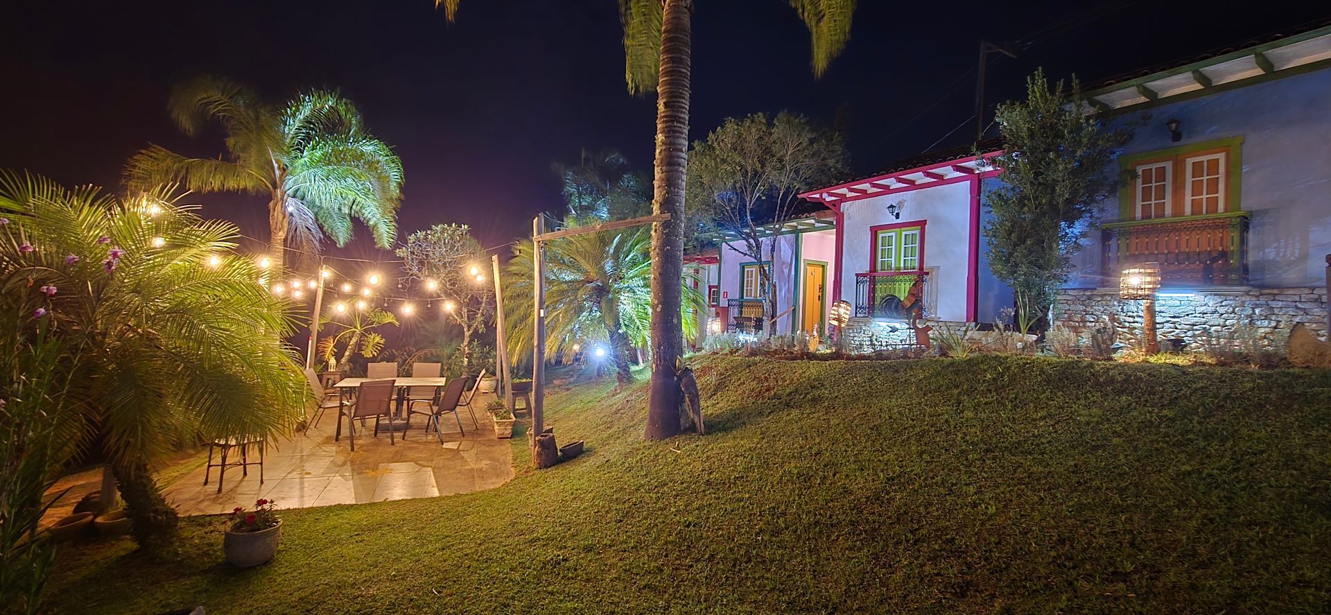 Jardim noturno colorido com luzes de corda, palmeiras e um caminho iluminado ao longo da casa.