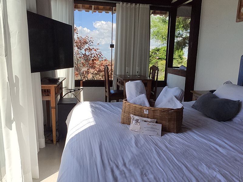 Quarto com cama arrumada, TV de parede e luz solar entrando pelas janelas abertas com vista para as árvores.