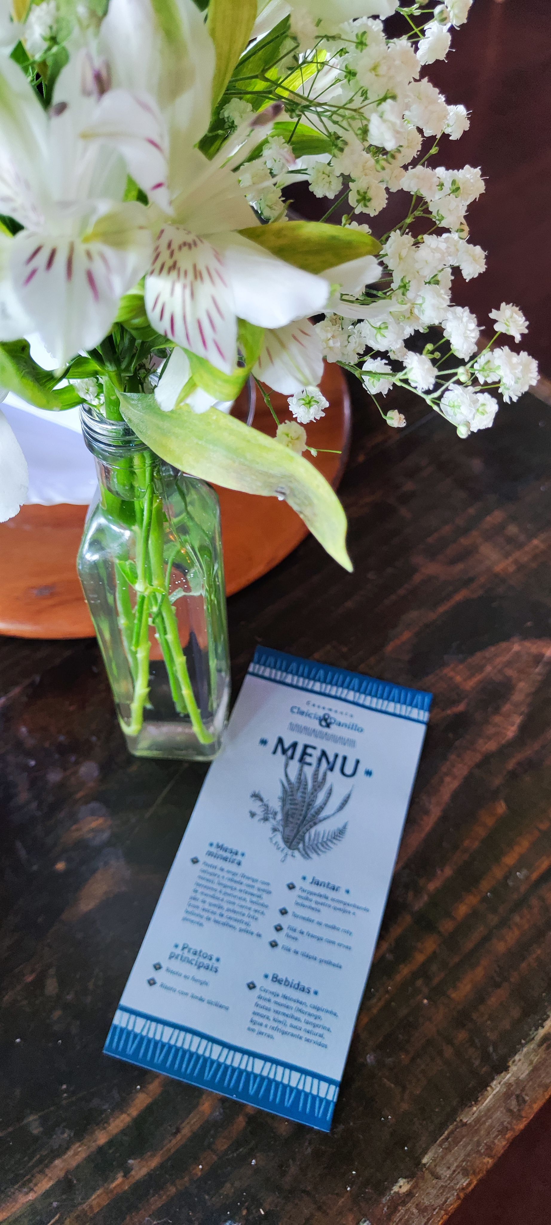 Flores brancas num vaso de vidro ao lado de um menu azul e branco sobre uma mesa de madeira.