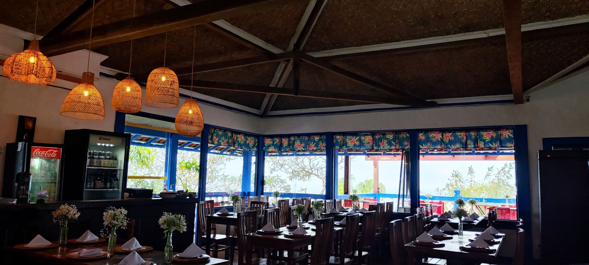 Interior de restaurante com iluminação suave, luminárias pendentes de vime e grandes janelas com vista para a água.