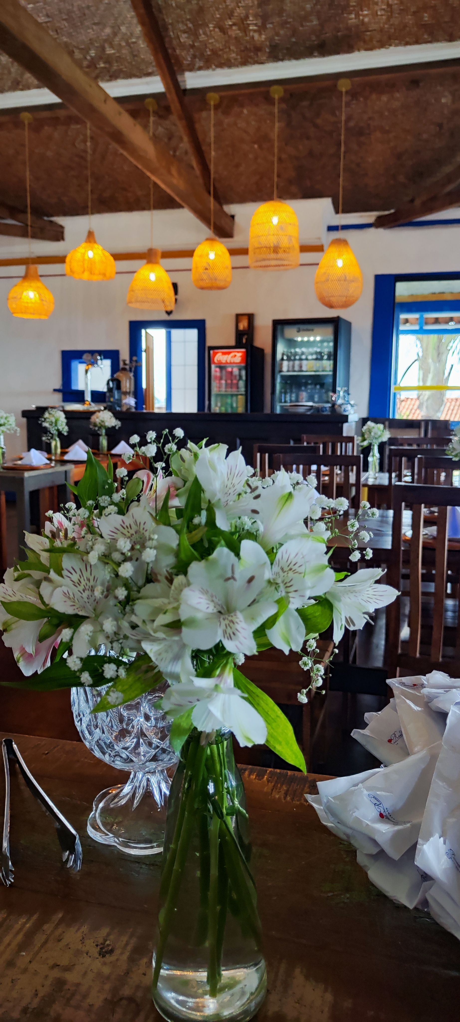 Interior de restaurante rústico com luzes âmbar penduradas e um vaso de flores brancas sobre a mesa.