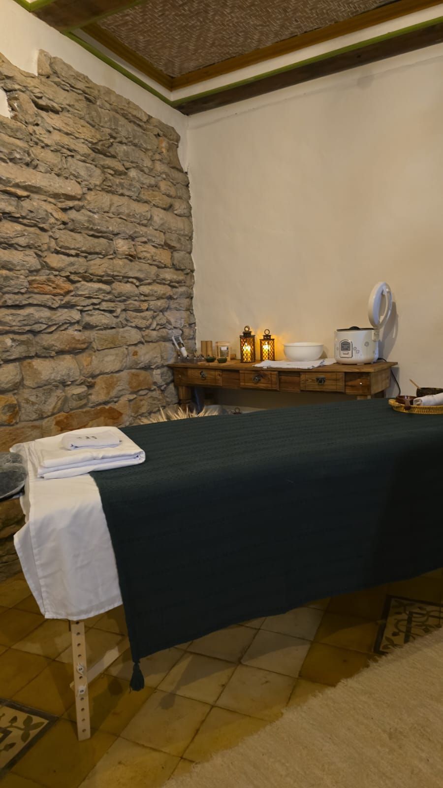 Mesa de massagem em uma pequena sala de spa com parede de pedra, toalhas, velas e decoração em uma prateleira de madeira.