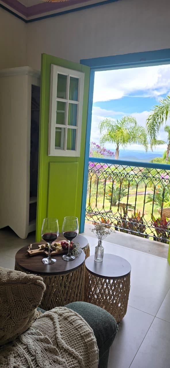 Sala de estar iluminada com porta verde aberta para uma varanda com vista para o mar e cadeiras de vime junto a uma pequena mesa.