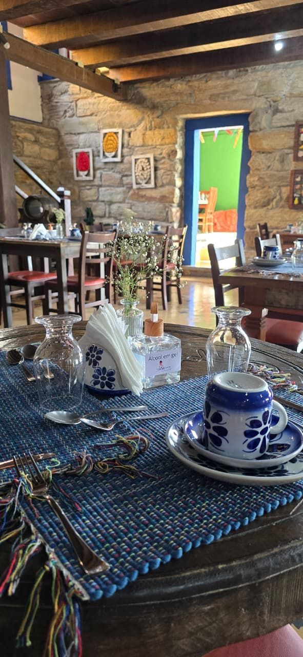 Mesa de café rústica com xícaras de estampa azul e jogos americanos de palha, paredes de pedra e uma porta iluminada ao fundo.