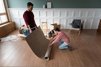 Couple rolls out a beige rug on a hardwood floor in a room with boxes and furniture; moving.