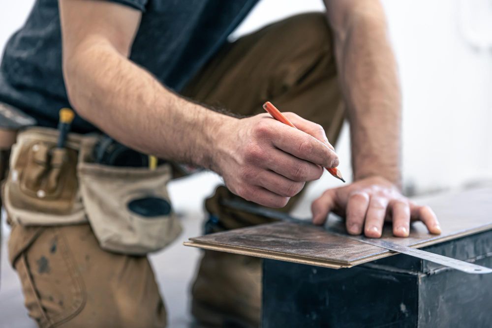 Person kneeling, using a pencil to mark a piece of wood for flooring installation.
