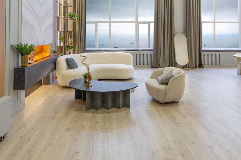 Living room with cream-colored couch, armchair, and coffee table. Light wooden floor, large windows, and fireplace.