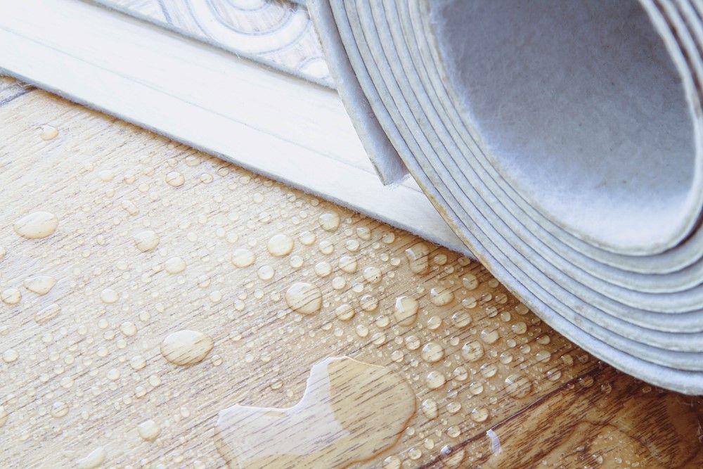 Water droplets on a light wood floor with a rolled-up gray vinyl flooring sample.