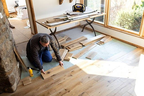 Man installing hardwood floor planks in a sunlit room, with tools on a table.