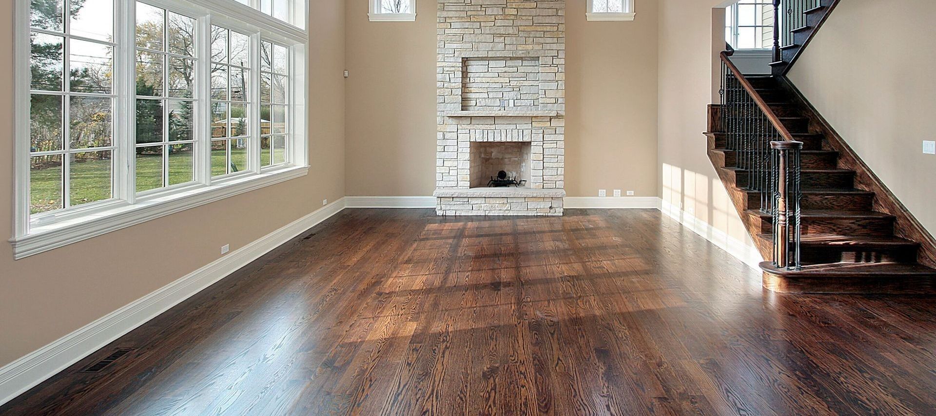 A living room with hardwood floors, a stone fireplace, and a staircase. Large windows let in sunlight.