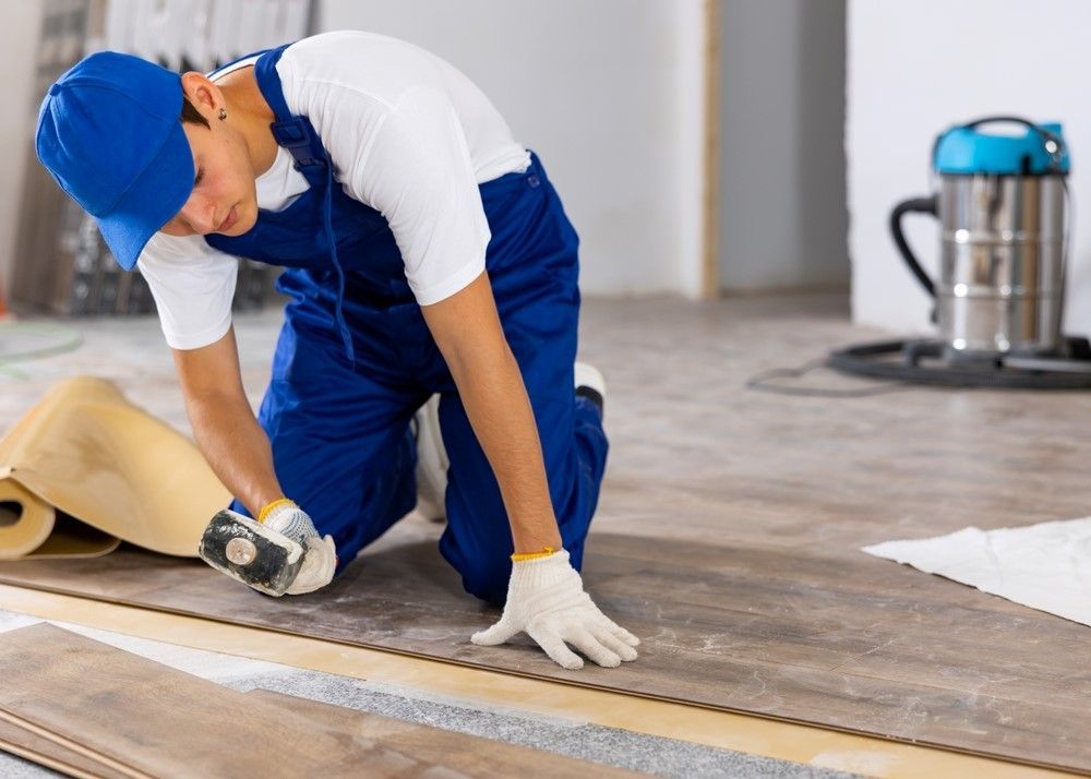 Person installing flooring, kneeling, wearing blue overalls, gloves, cap, vacuum in background.