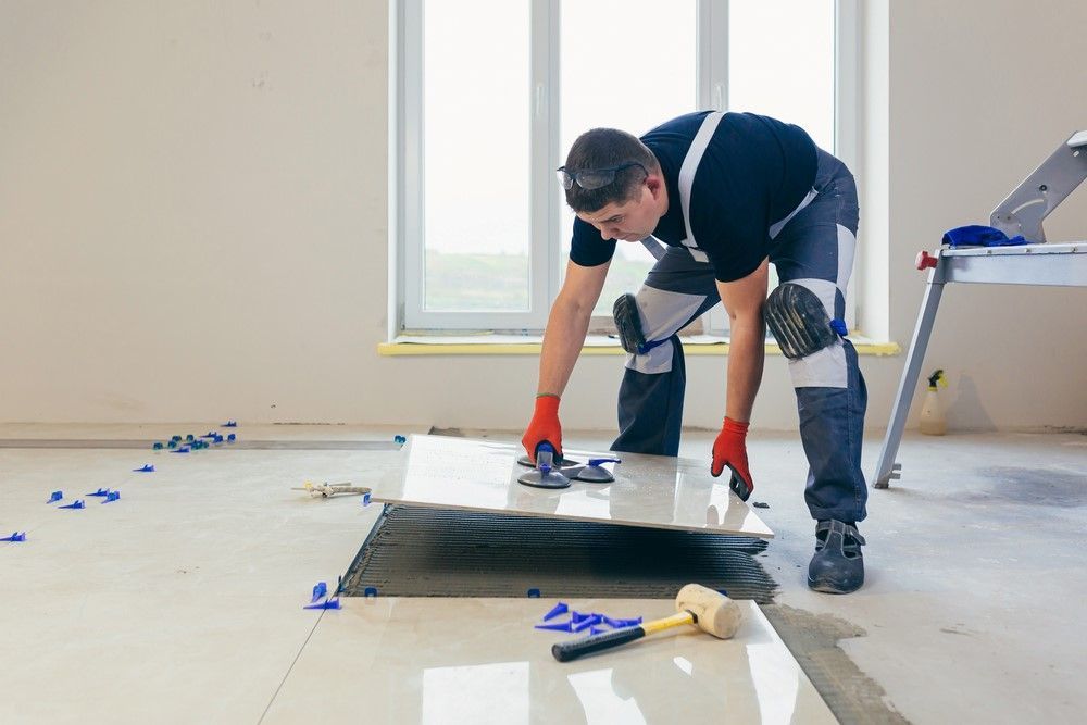 Man tiling a floor, kneeling and using tools in a room with a window, grout spread on the tiles.