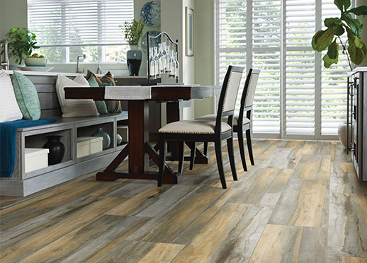 Dining room with a wooden table, built-in bench, and wooden-look floor. Sunlight streams in from the window.