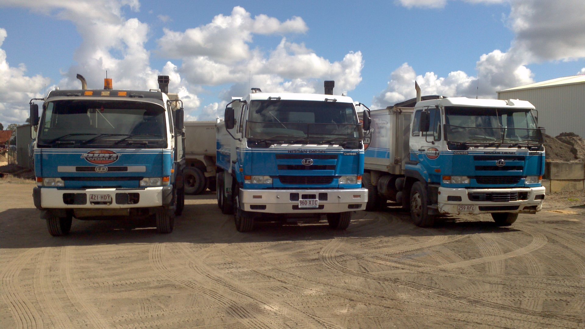 Three blue trucks parked side by side on a dirt lot under a cloudy sky