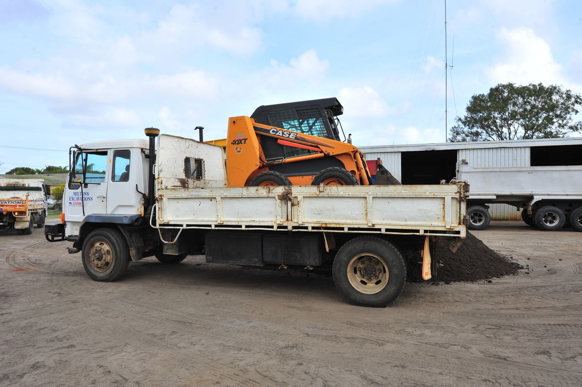 White flatbed truck loaded with an orange skid steer on a gravel lot
