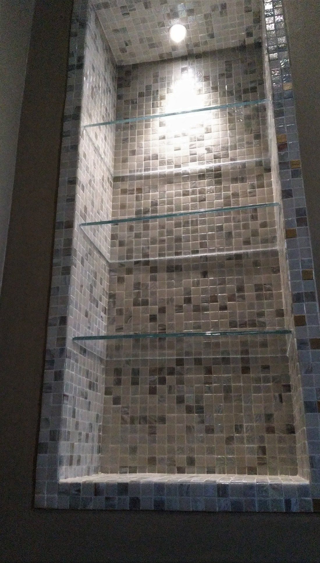 Una nicchia doccia incassata con piastrelle a mosaico beige, tre mensole in vetro e una luce a soffitto.