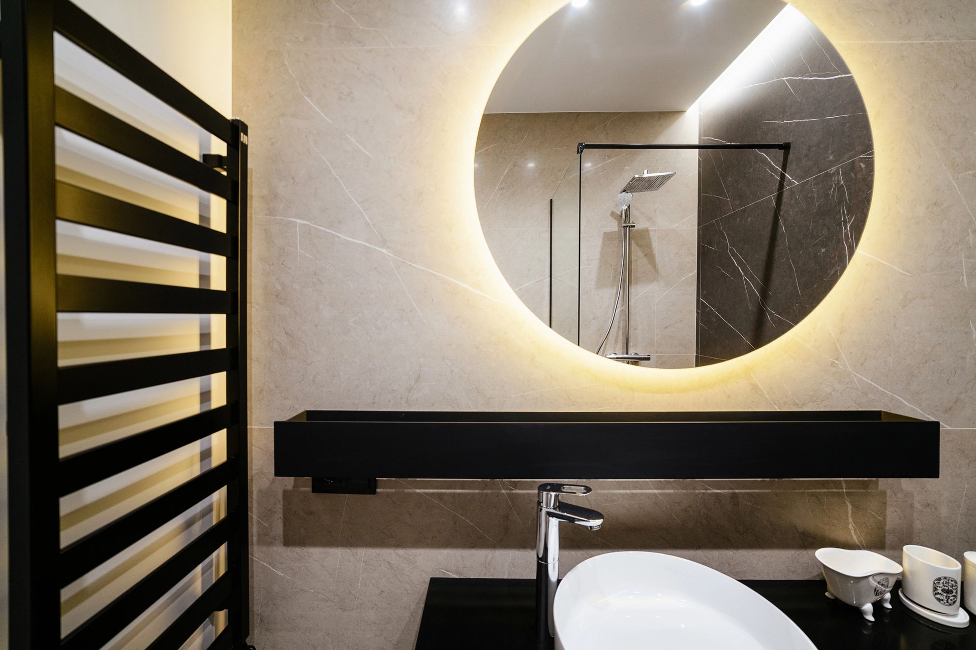 Un bagno moderno è caratterizzato da uno specchio rotondo retroilluminato, un mobile sospeso nero con lavabo a ciotola bianco e un portasciugamani nero.
