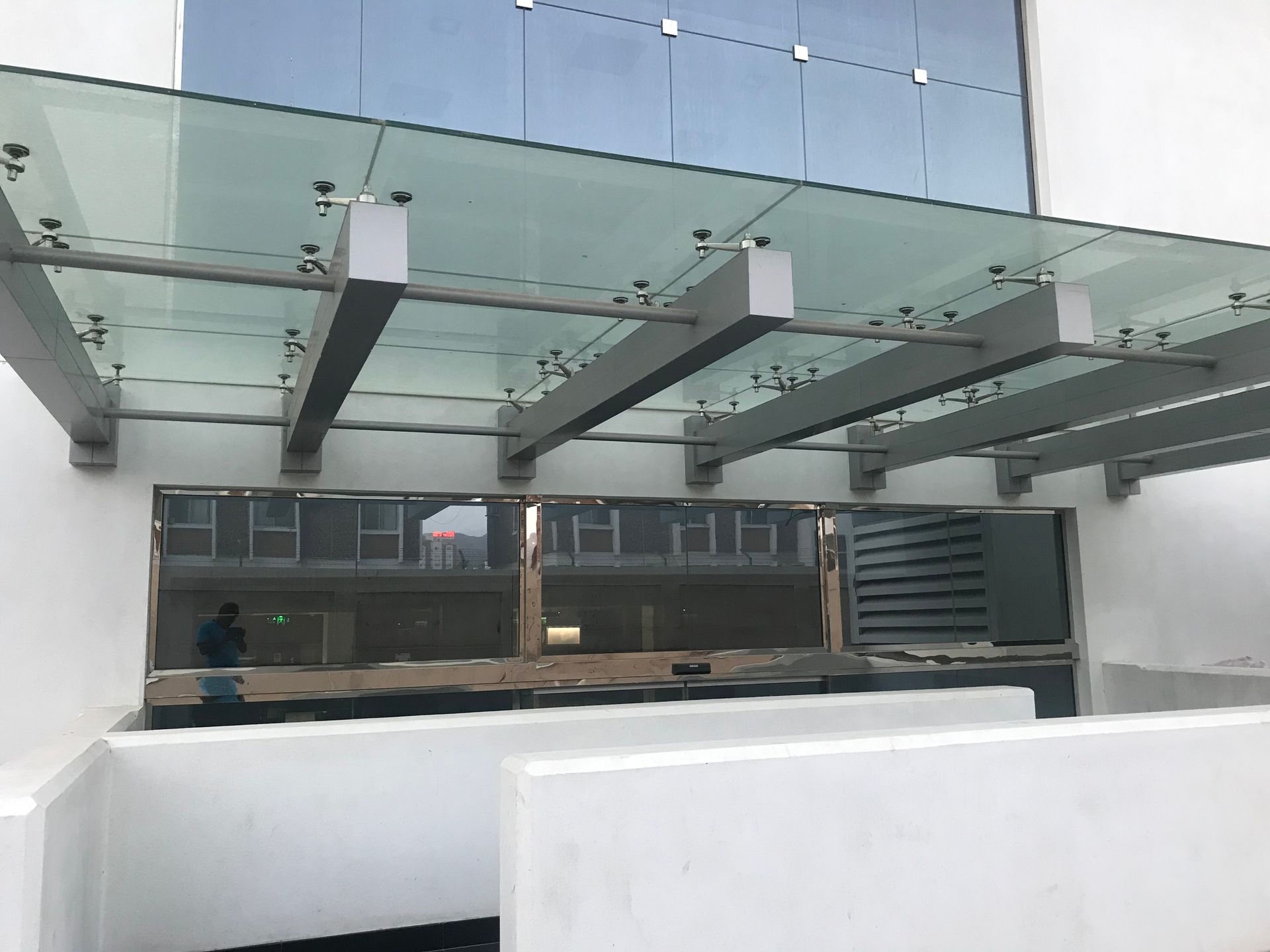 Una tettoia in vetro con supporti metallici si estende sopra l'ingresso di un edificio con pareti bianche e una vetrina in vetro riflettente.