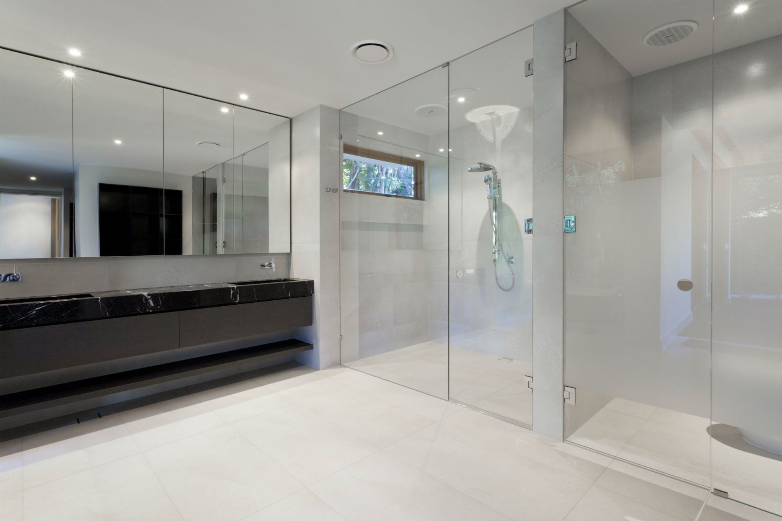 Bagno moderno con un lungo mobile lavabo nero, grandi specchi e una cabina doccia con pareti in vetro su un pavimento piastrellato di bianco.