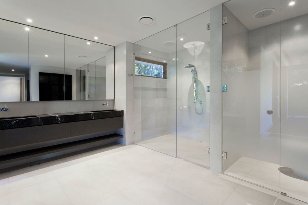 Bagno moderno con un lungo mobile lavabo nero, grandi specchi e una cabina doccia con pareti in vetro su un pavimento piastrellato di bianco.