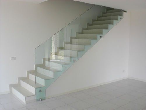 Scala moderna con gradini bianchi e ringhiera in vetro senza telaio montata lateralmente su una parete bianca liscia.