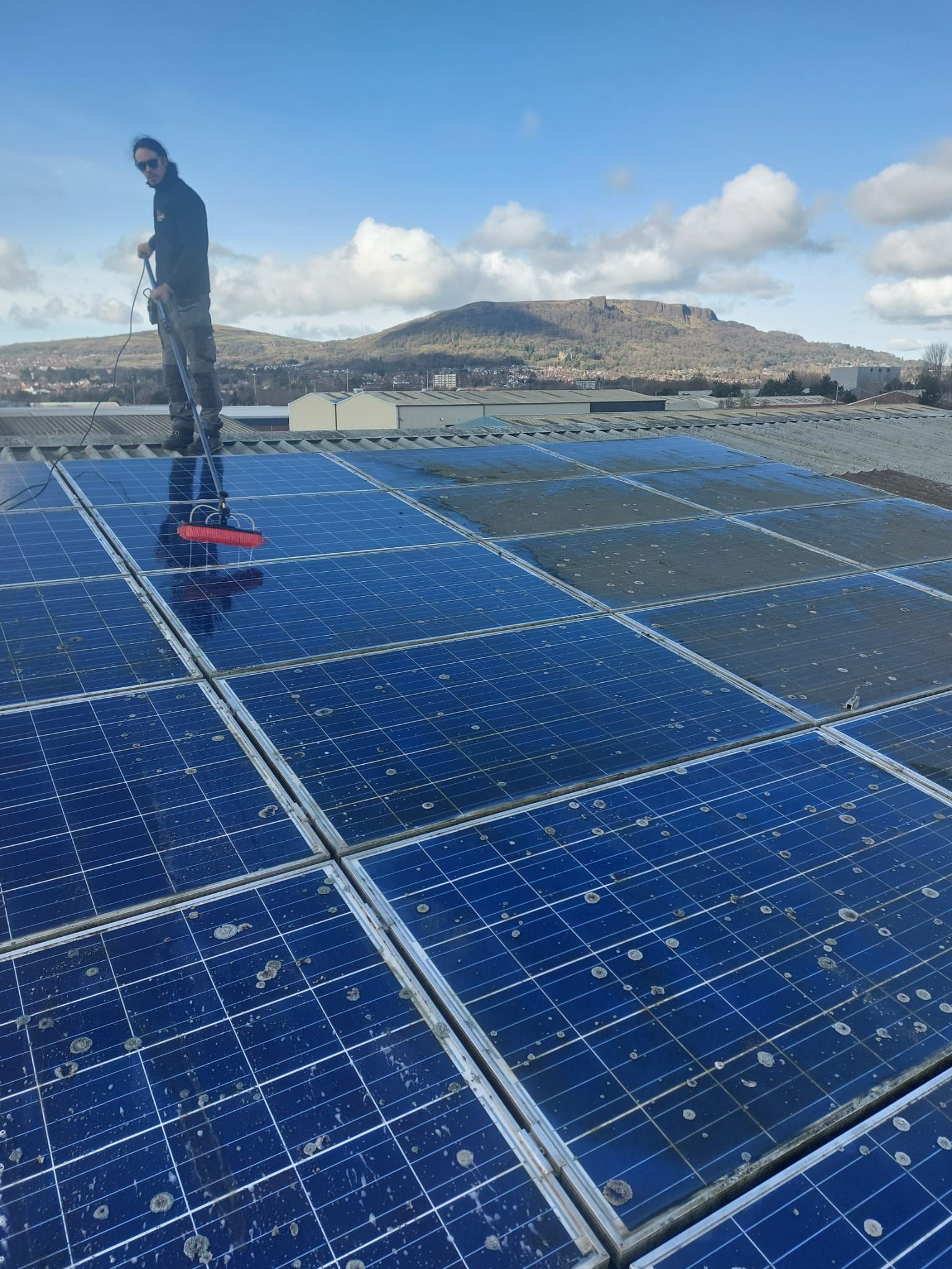 A man is standing on top of a roof cleaning solar panels.