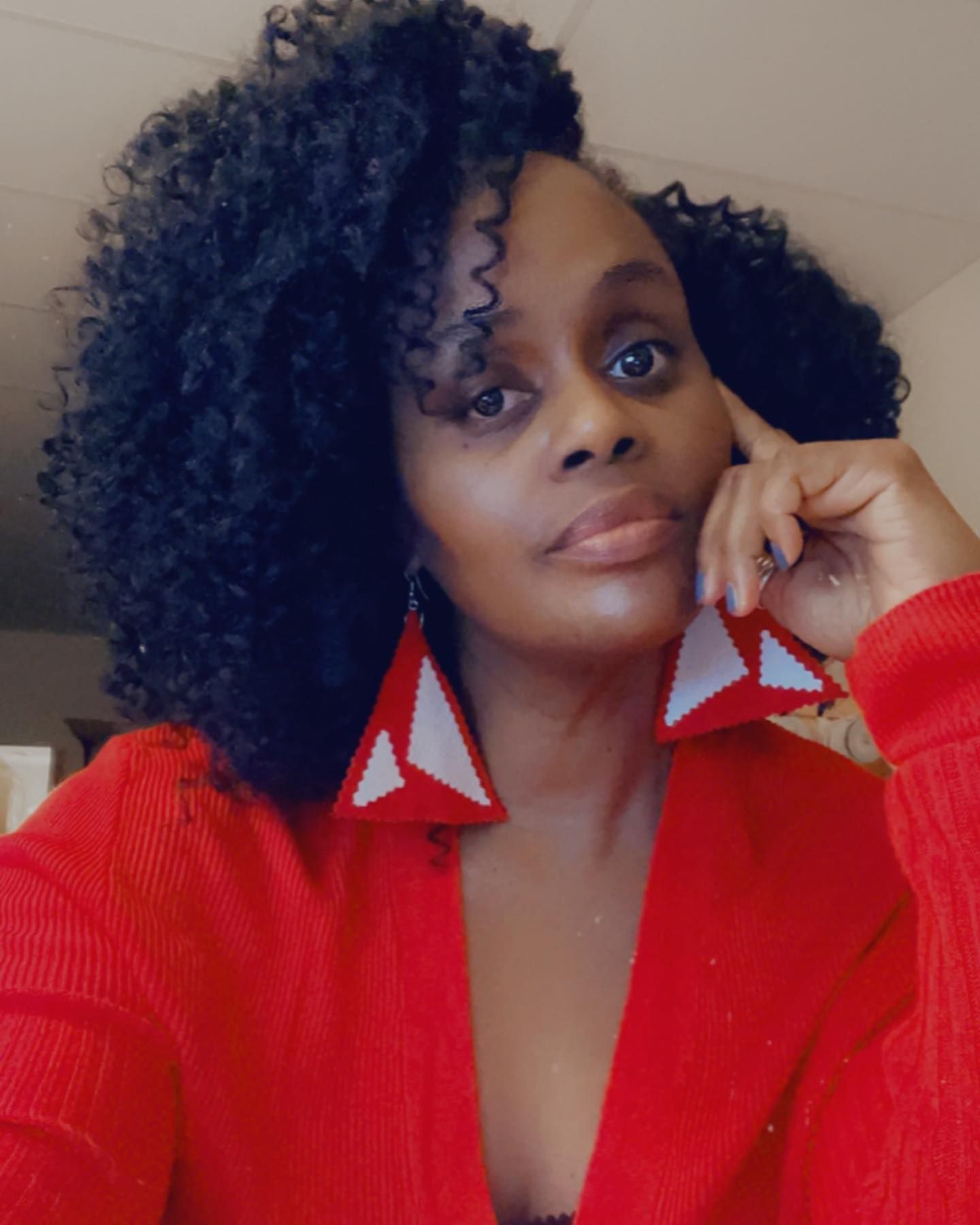 Woman with dark curly hair wearing red sweater and large red and white triangle earrings, hand on face.