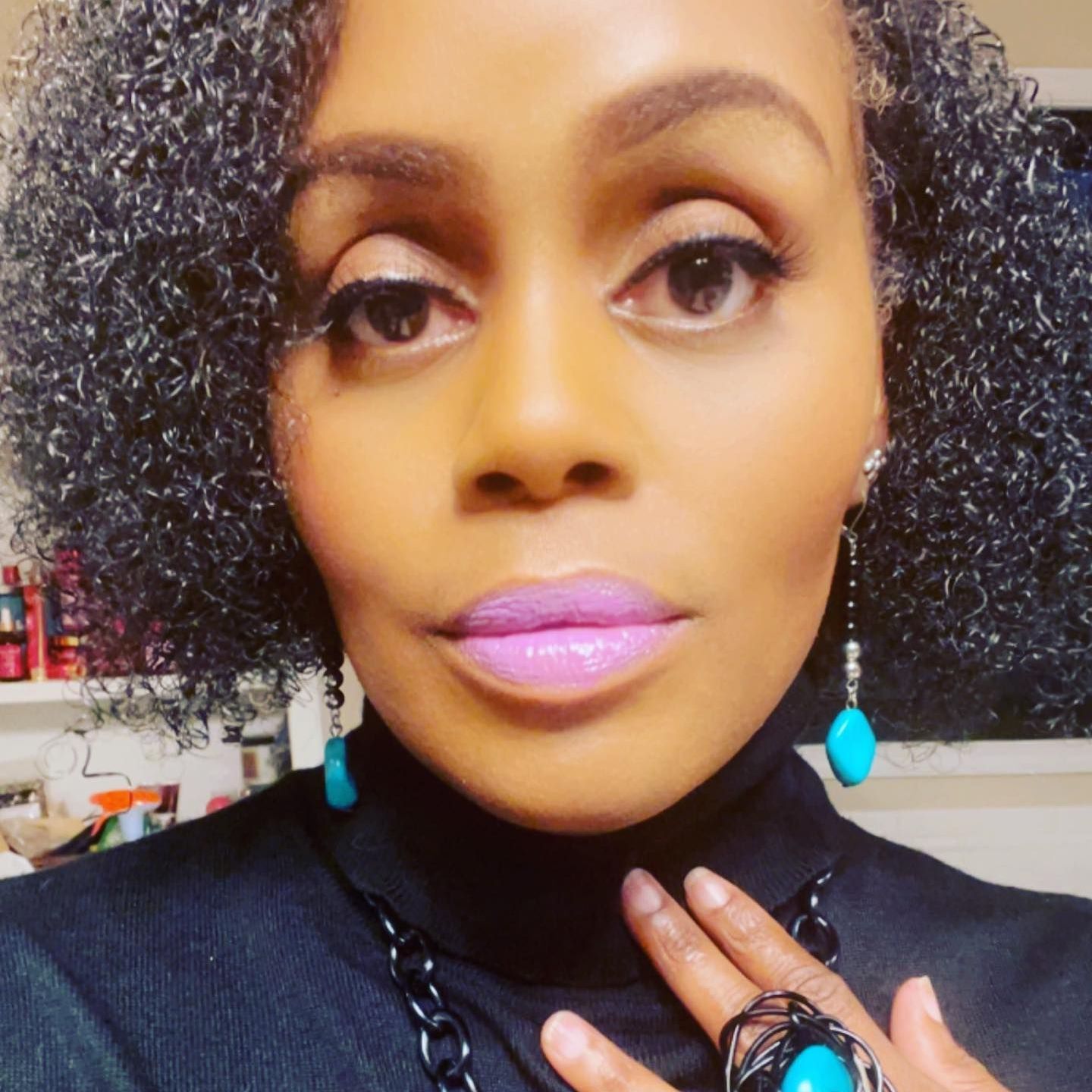 Woman with curly gray hair, wearing turquoise jewelry and a black turtleneck, pink lipstick.