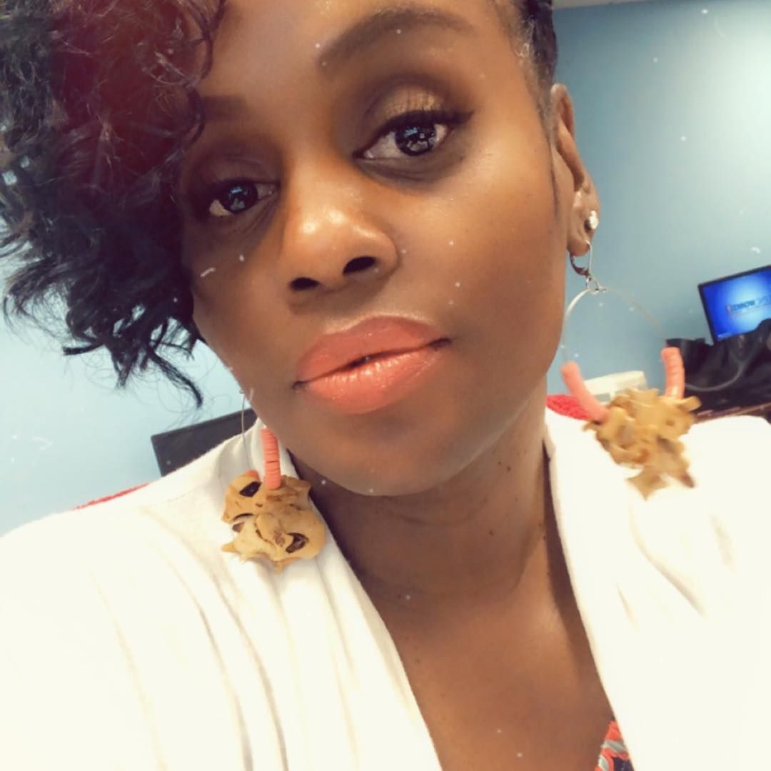 Woman with curly hair, wearing light lipstick and floral earrings, in an office setting.