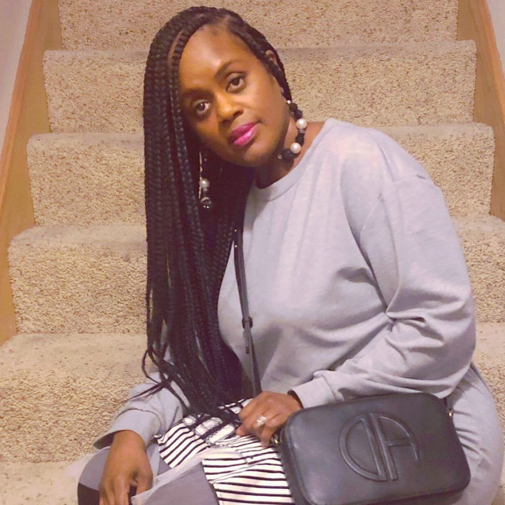 Woman with long braids, wearing a gray outfit and purse, seated on carpeted stairs.