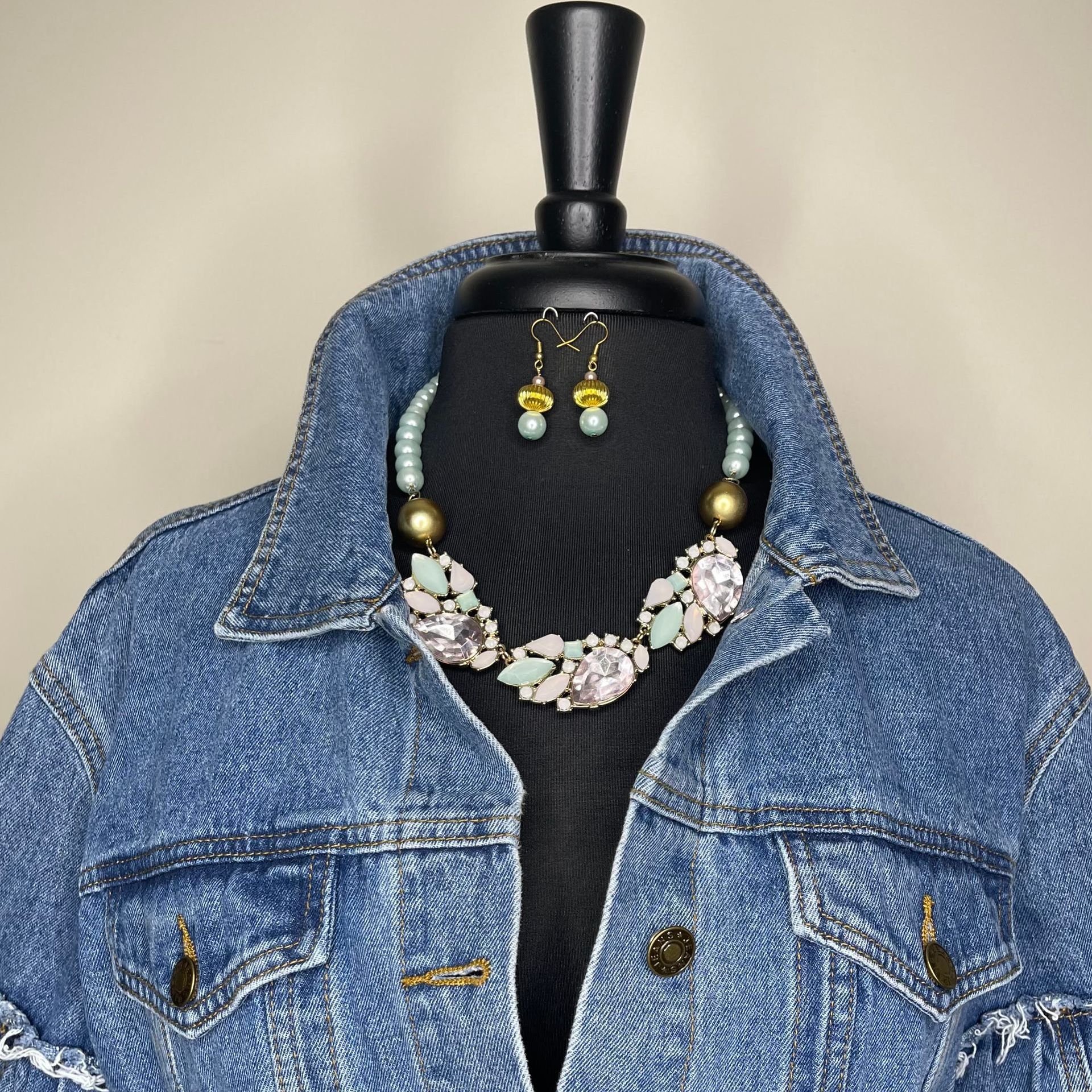 Denim jacket with necklace and earring set on a black mannequin. Gold accents, pastel gems, and blue beads.