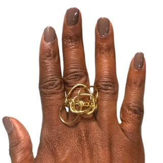 A person's hand with brown nails wearing a gold intertwined ring, set against a white background.