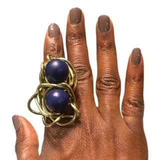 Hand wearing a large gold ring with two dark blue orbs. The hand has brown nails.