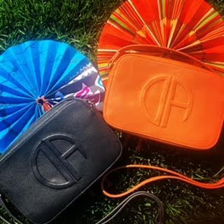Two Telfar bags: black and orange, with blue and orange paper fans on grass.